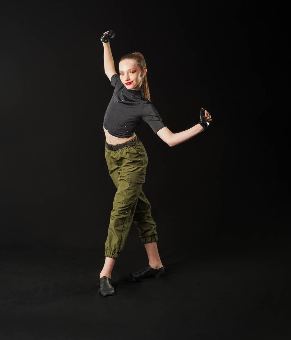 A dancer in cargo pants and crop top performs an energetic move against a black backdrop.