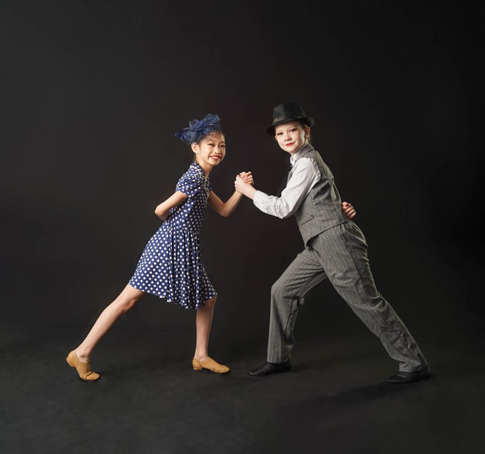Two dancers perform a vintage-style dance scene in period costumes against a dark background.