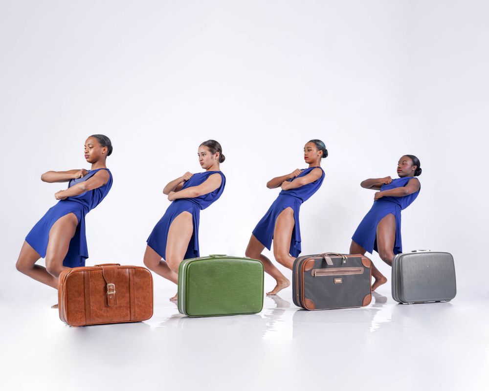 Four dancers in blue dresses pose identically leaning against vintage suitcases in a line.