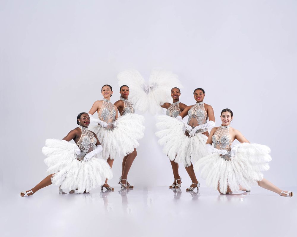 Dancers in white feathered costumes perform in formation against a light backdrop.