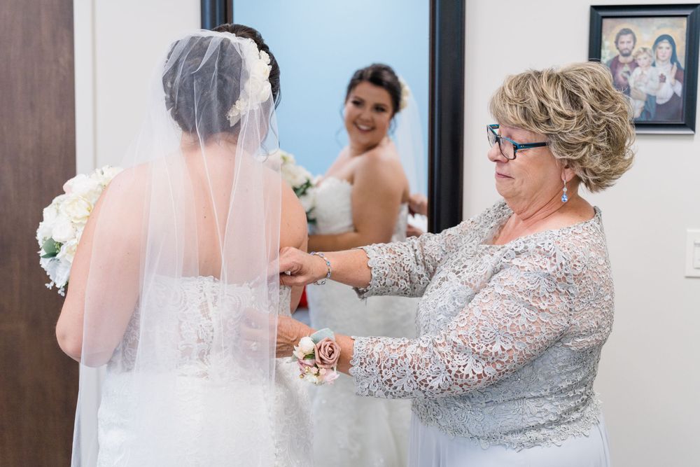A woman in a grey lace dress helps adjust a bride's white wedding gown and veil during preparation.