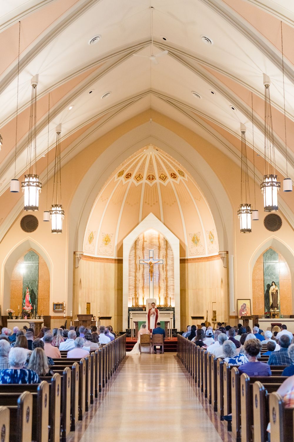 Elegant church interior with vaulted ceilings, hanging lanterns, and cream-colored walls filled with wedding guests.