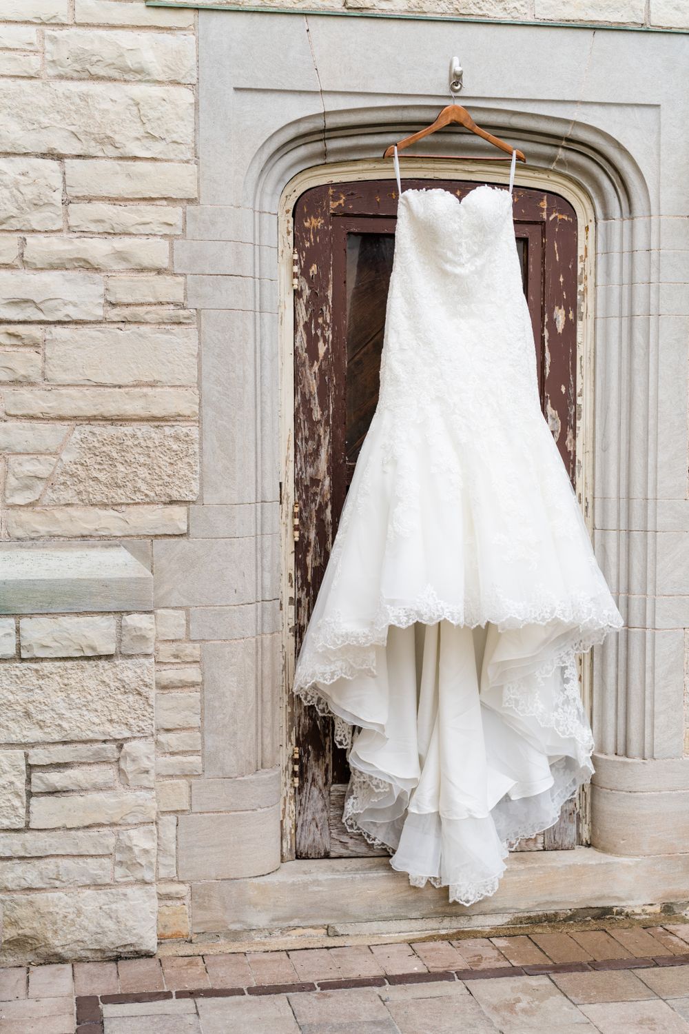 An elegant white lace wedding dress hangs on a wooden hanger against a stone archway.