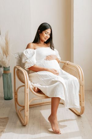 Person in white maternity dress sitting gracefully in rattan chair with natural decor.