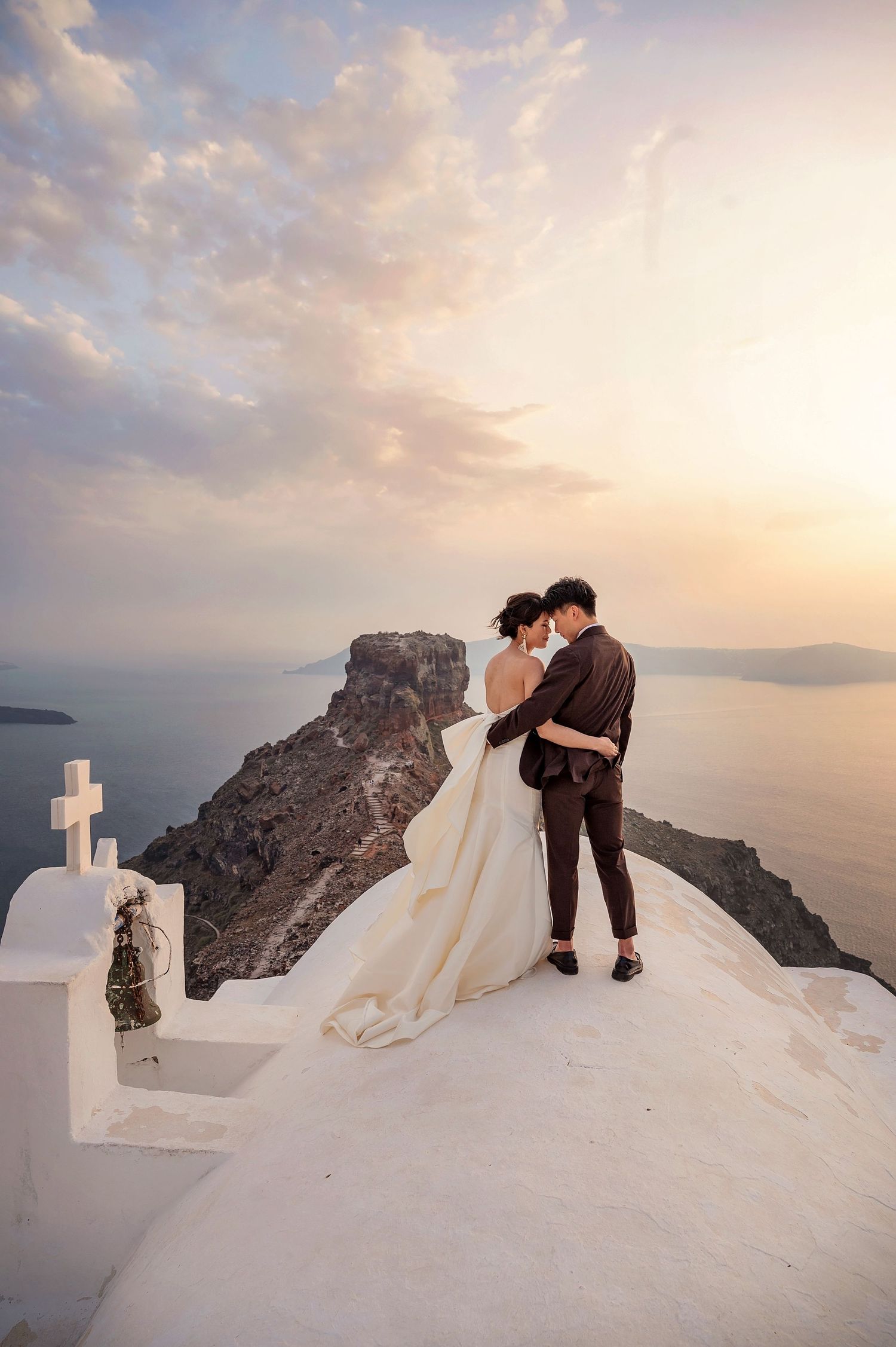Wedding couple embracing on cliff edge with dramatic Santorini sunset view.