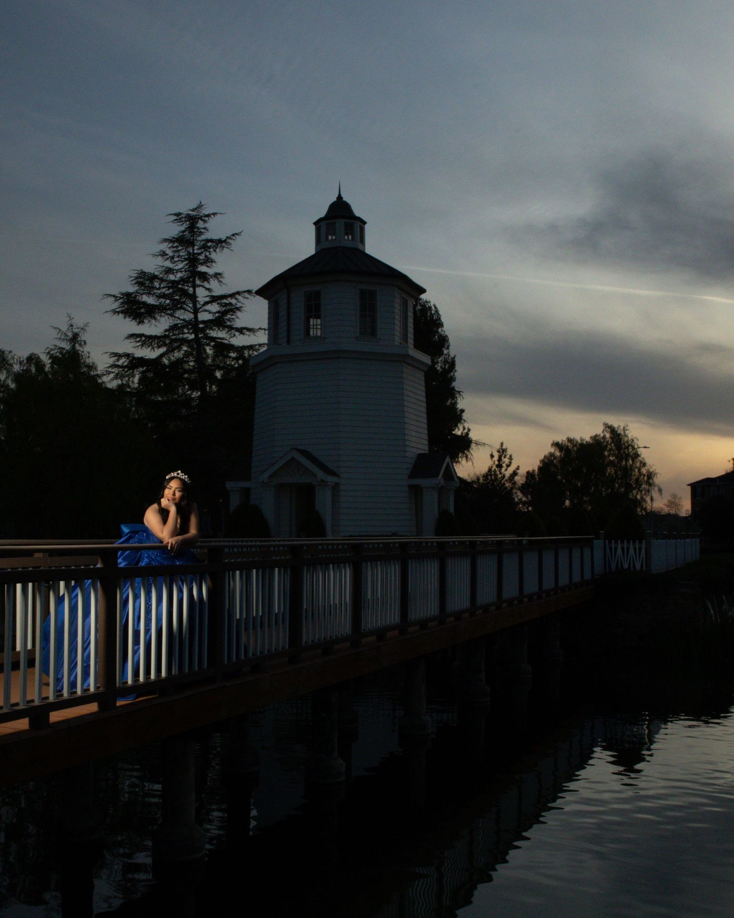 A silhouetted lighthouse and boardwalk at dusk with a person sitting along the railing beside calm reflective waters.