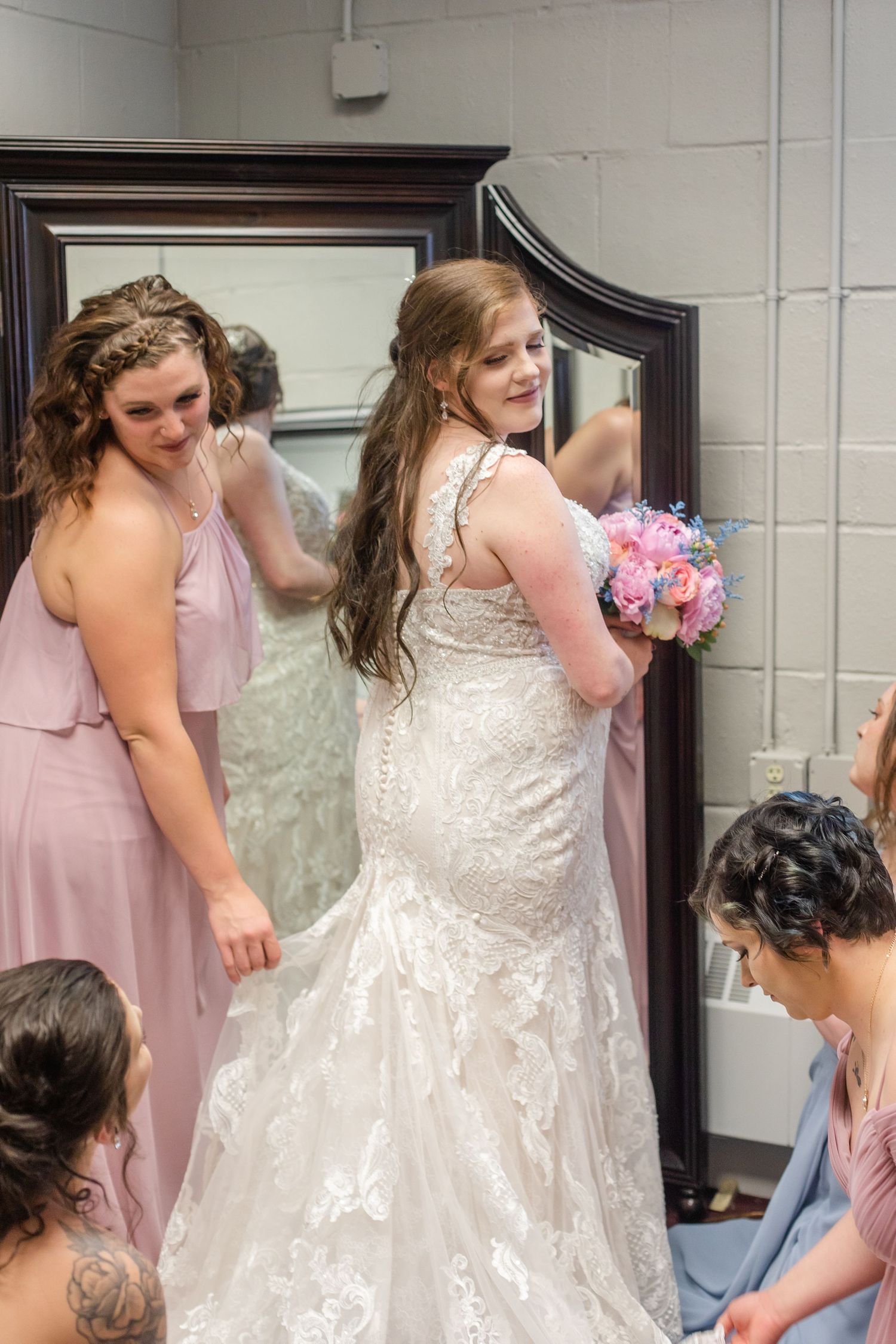 A bride in a lace wedding gown stands before a mirror while bridesmaids in pink dresses assist her.