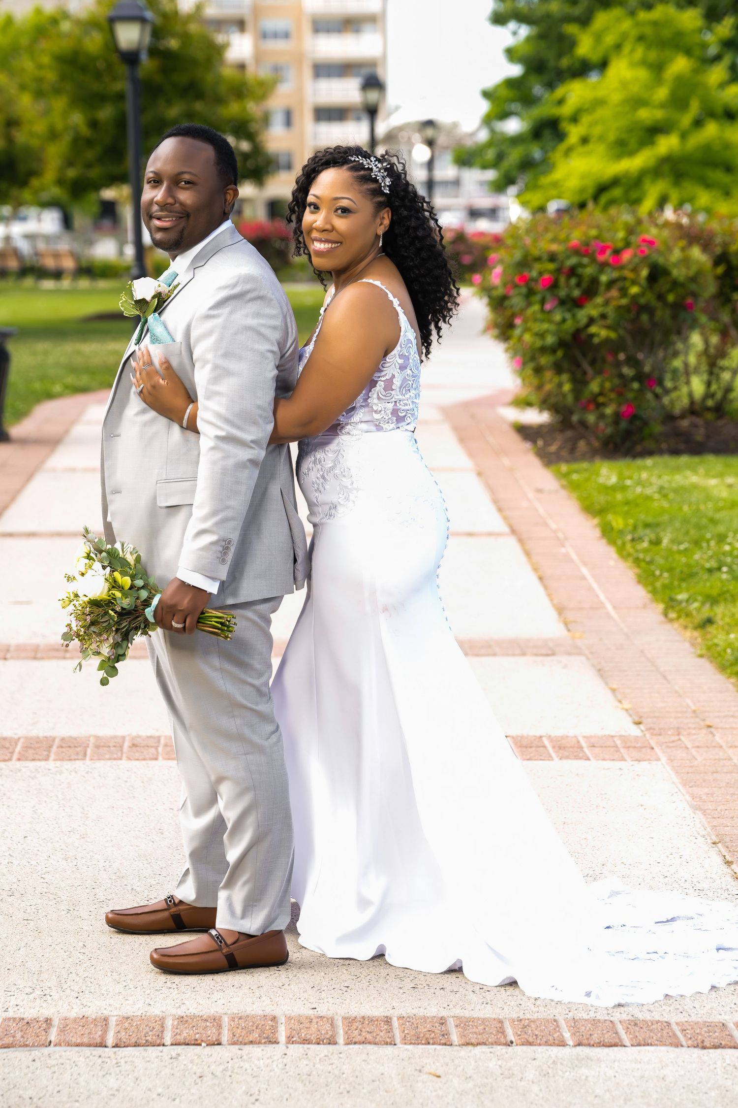 A wedding couple poses back-to-back on a brick pathway with landscaping in the background.