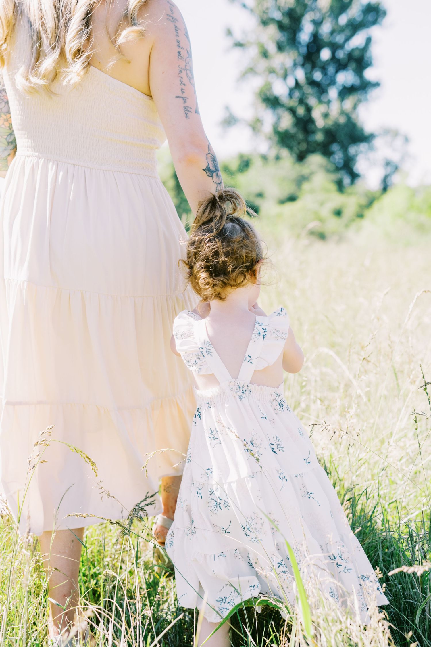 A mother and child in white dresses hold hands while walking through a sunny meadow.
