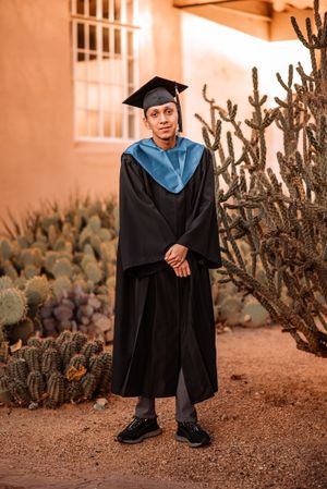 A graduation photo featuring a graduate in cap and gown posing next to desert cacti and a stucco building.