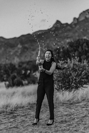 Black and white photo of someone celebrating by popping champagne outdoors with mountains in background.