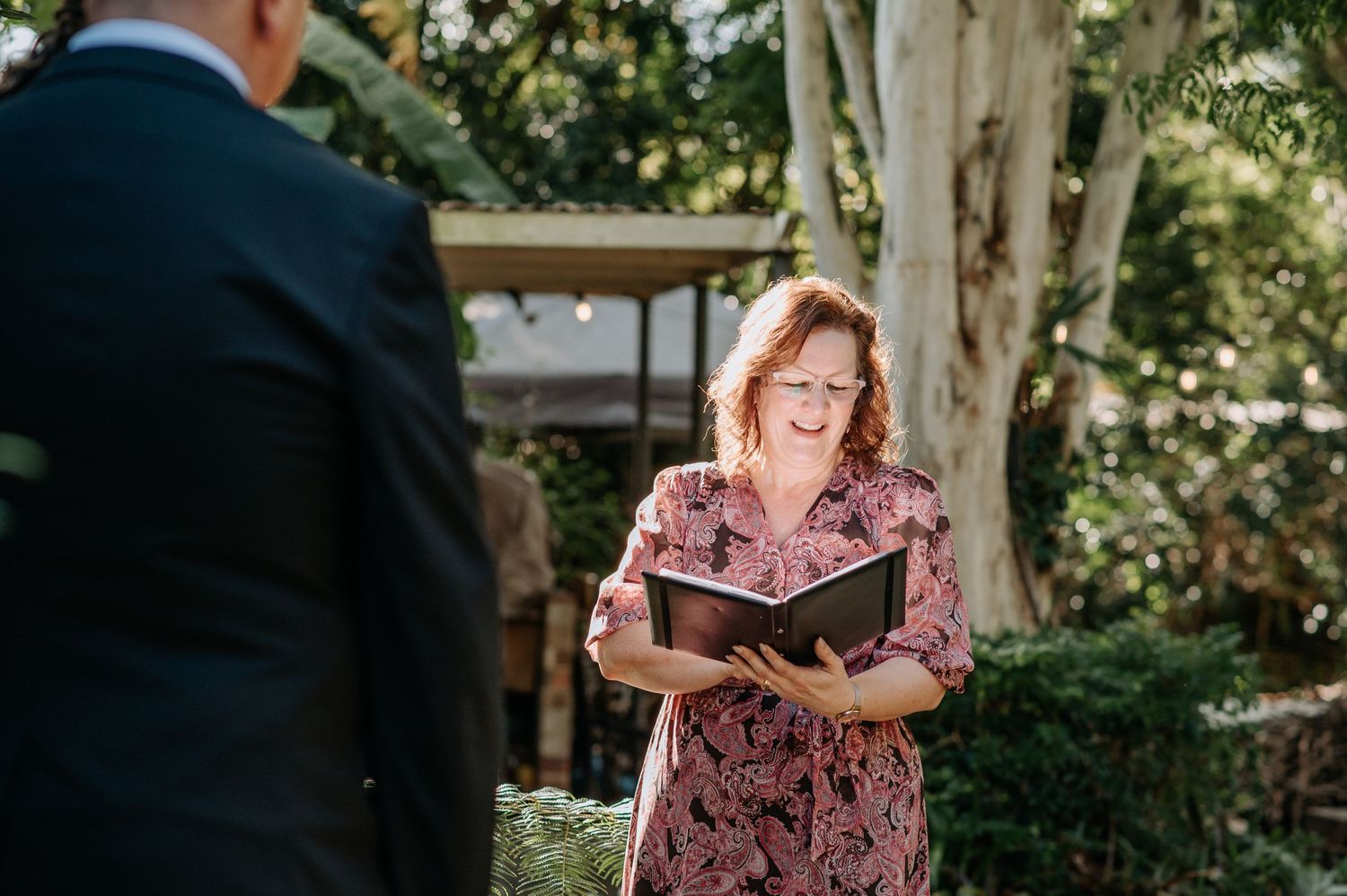 An officiant in a floral dress reads from a book during an outdoor ceremony.