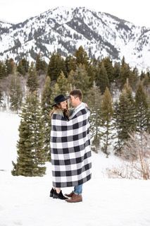 couple snuggling with a blanket at Snowbasin utah with pine trees and mountain in the background.