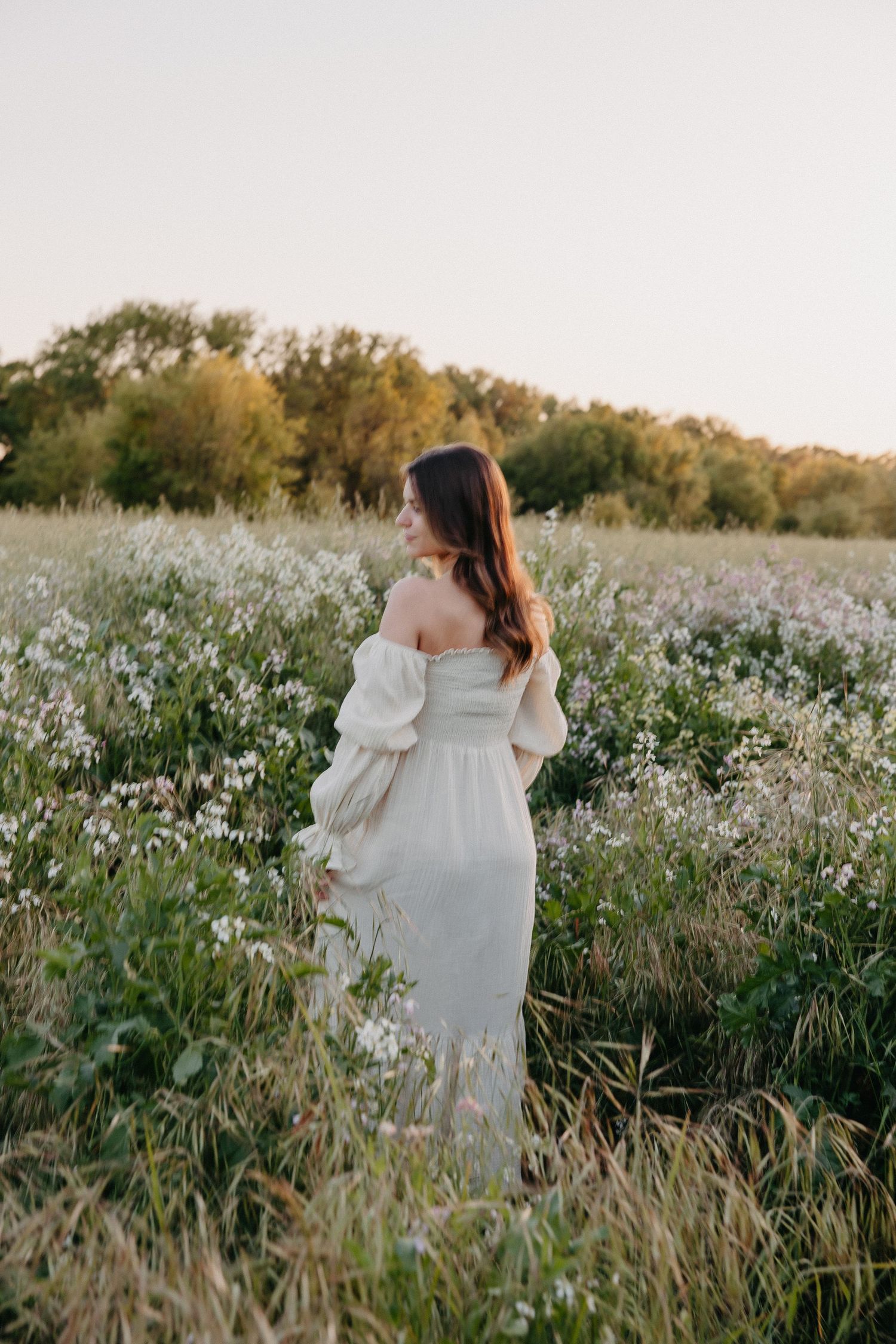 Woman in off-shoulder white dress stands among wildflowers in evening light.