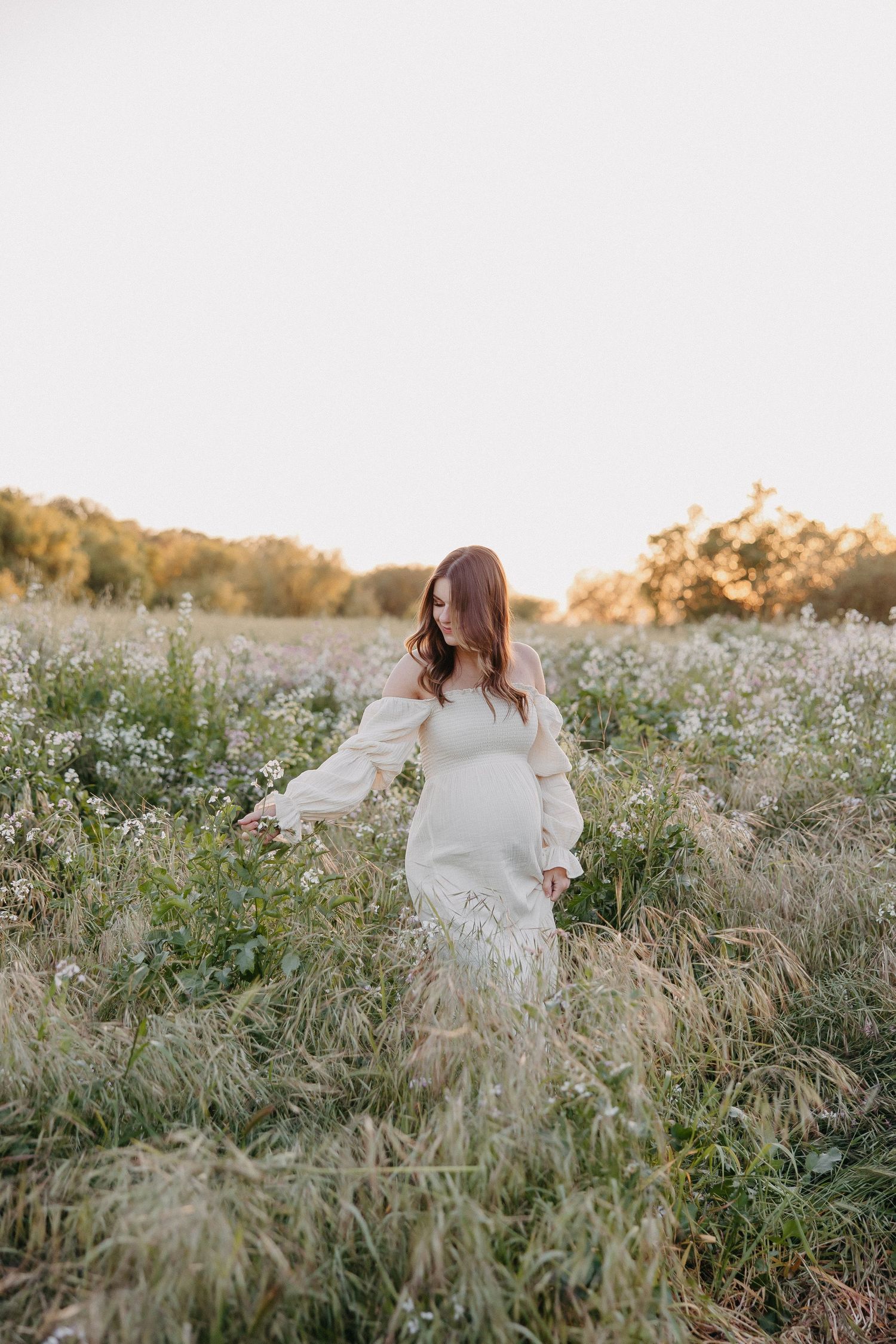 Side view of woman in white dress standing among tall grasses and wildflowers at sunset.