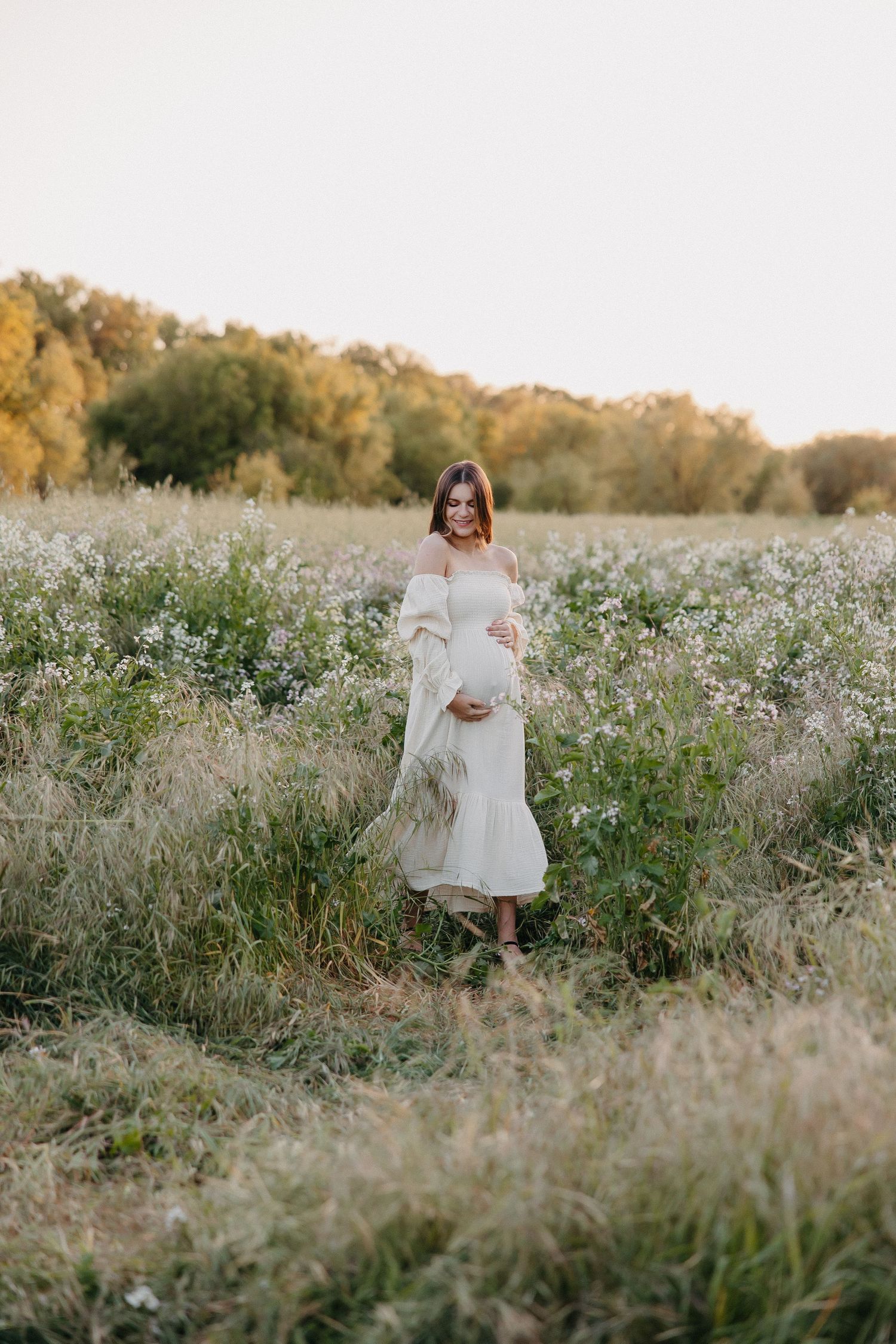 Expectant mother in white dress stands gracefully in field of autumn wildflowers.