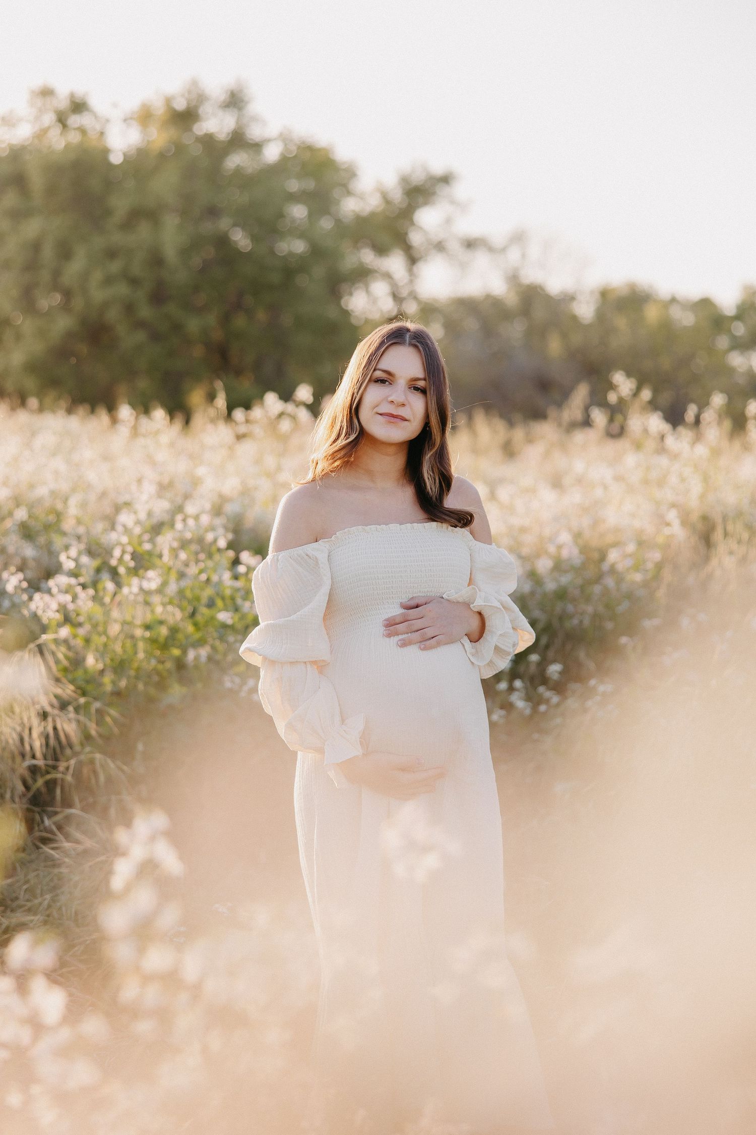 Maternity portrait of woman in white dress surrounded by sunlit wildflowers.