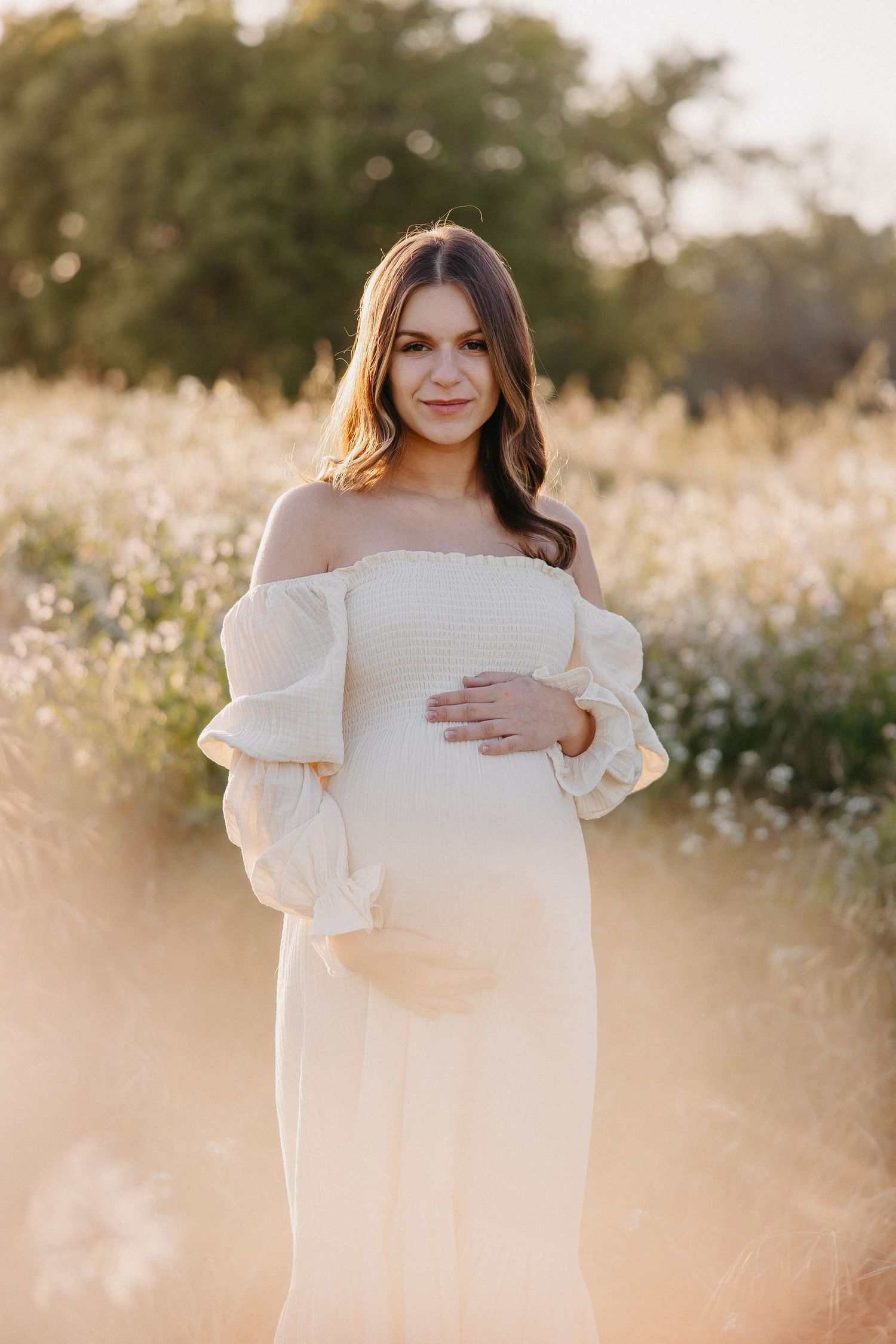 Glowing expectant mother in off-shoulder white dress poses in golden sunset field.