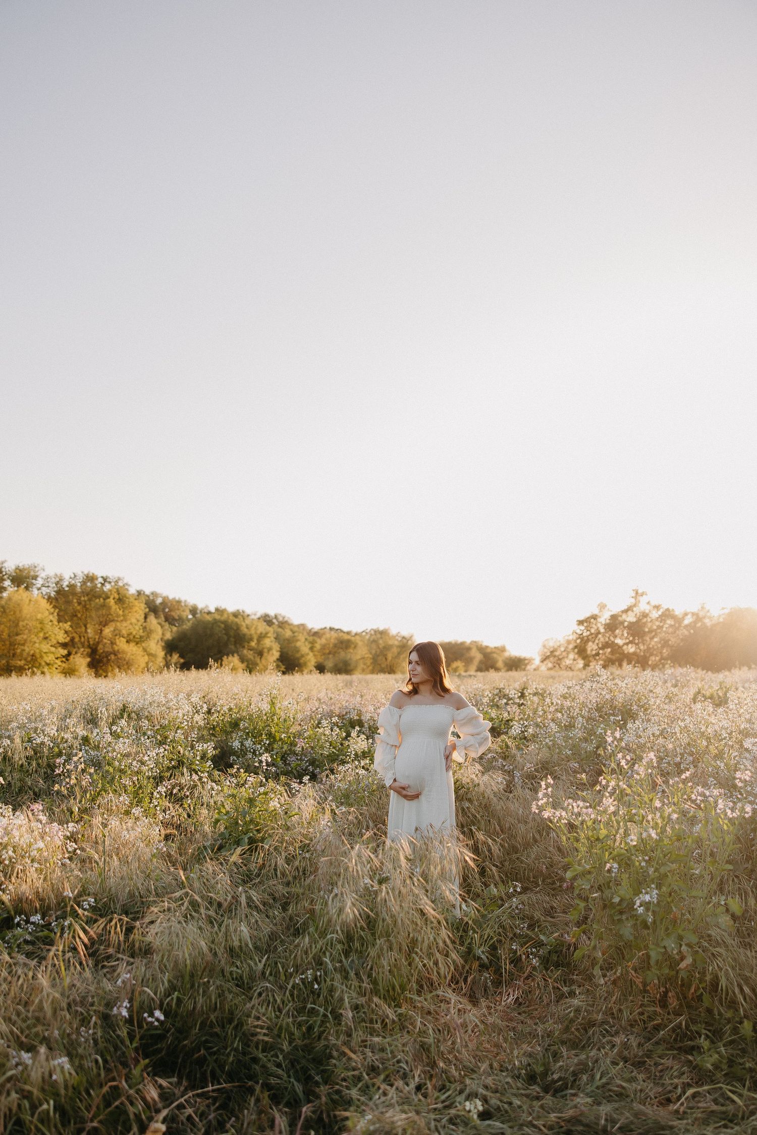 Ethereal maternity scene in sunlit meadow with white dress against autumn trees.