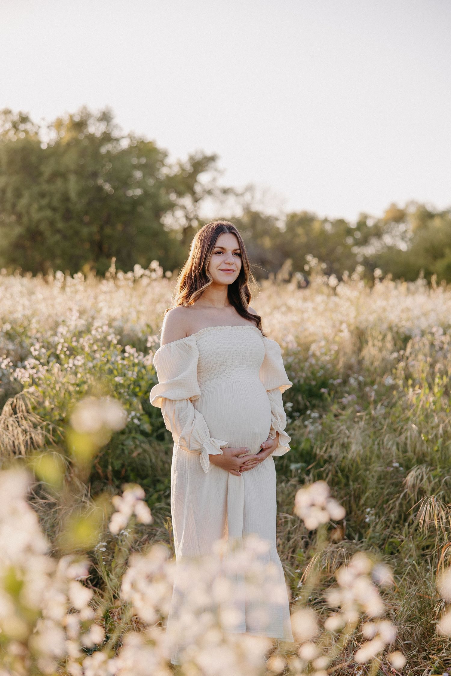 Expectant mother in white off-shoulder dress poses in wildflower field during golden hour.