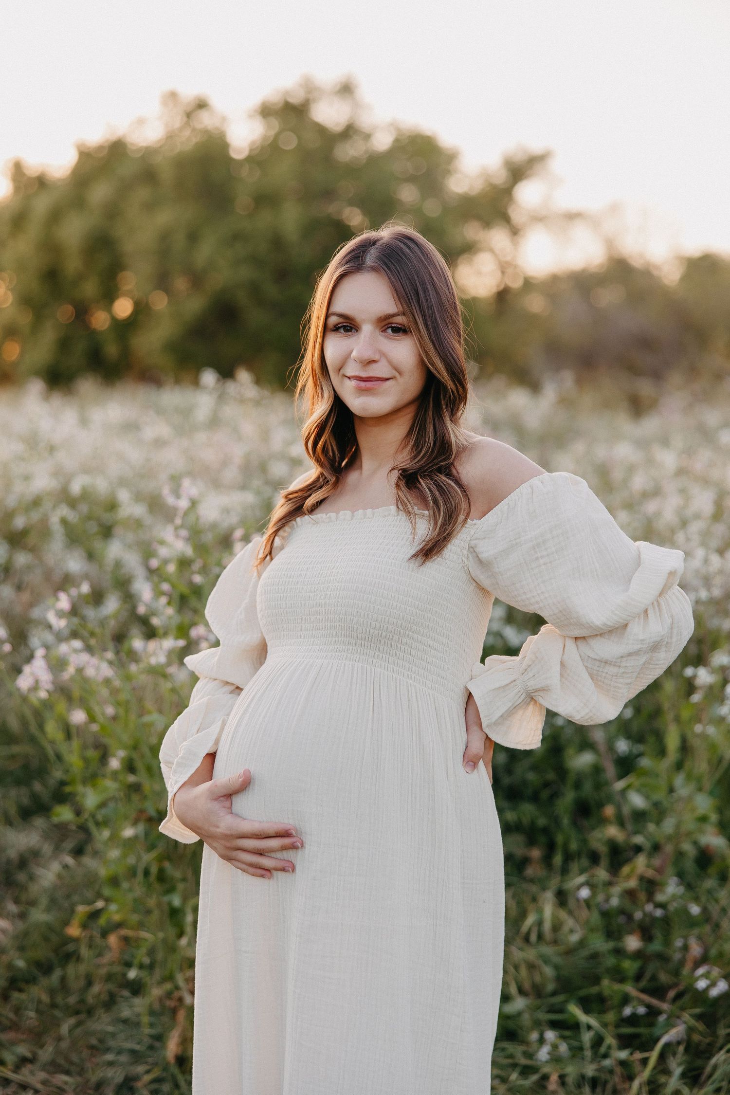Sunset maternity photo shows woman in flowing white dress cradling baby bump in field of wildflowers.