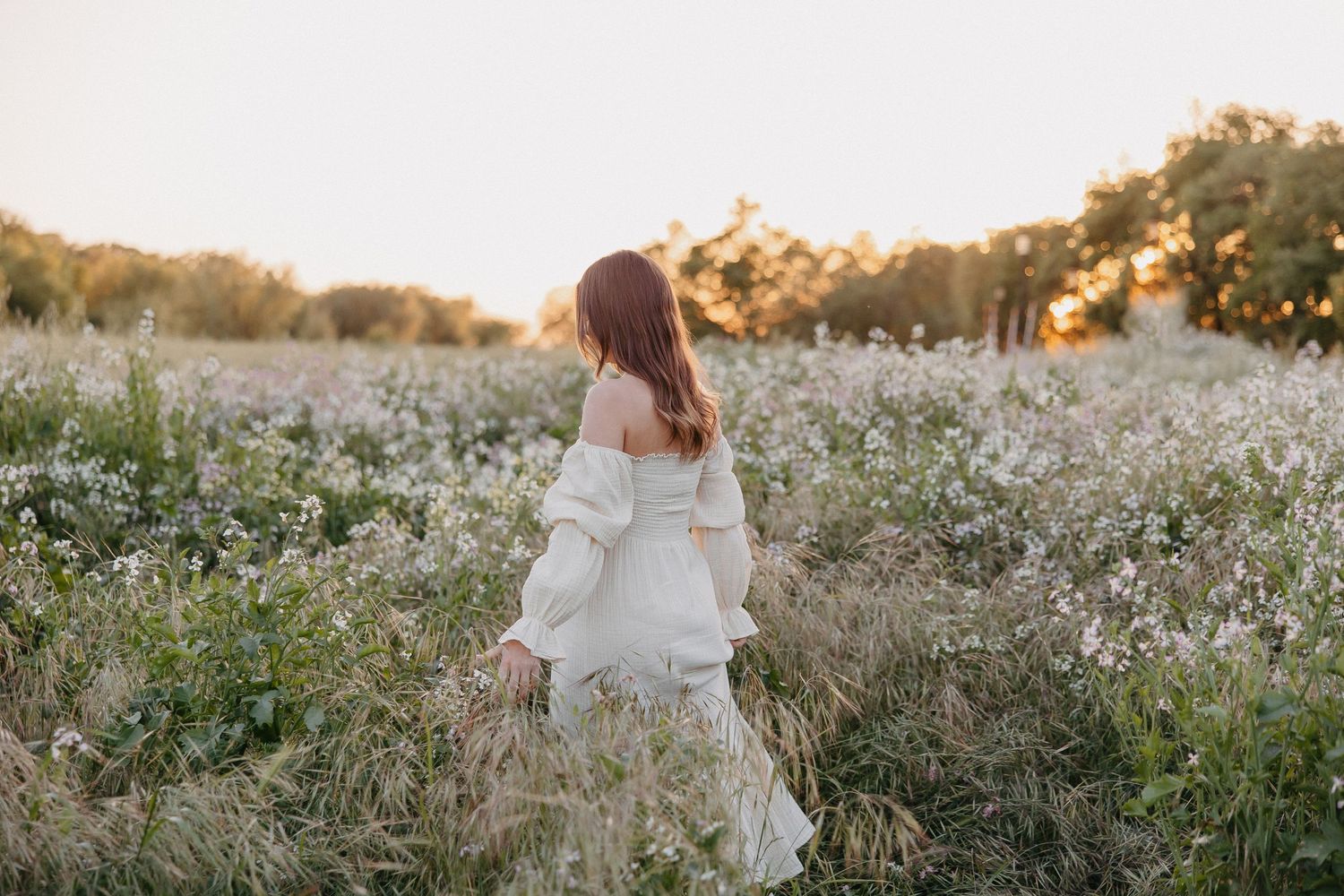 Woman in romantic white dress walking through meadow of white flowers at dusk.