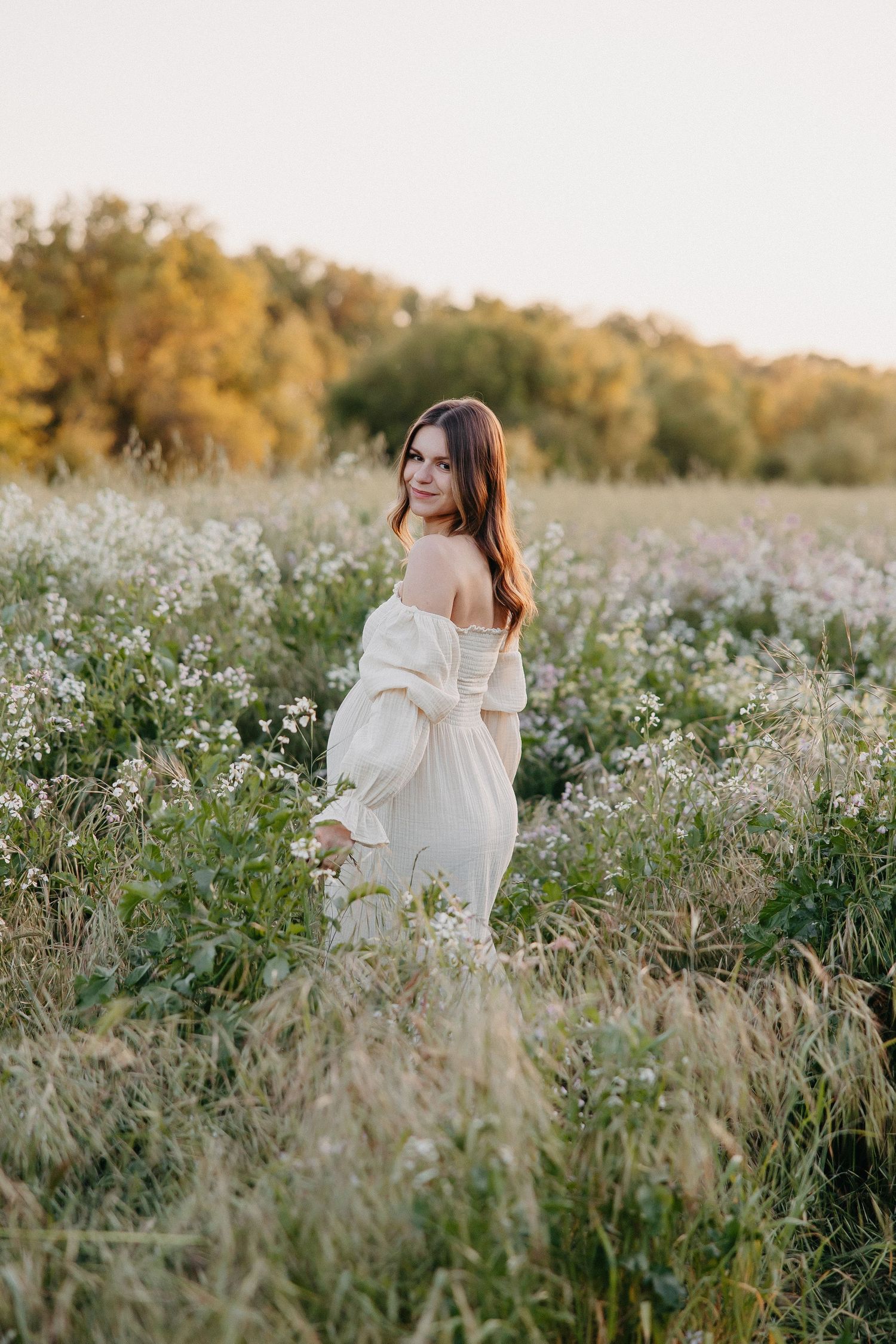 Woman in flowing white dress poses amid field of wildflowers during golden hour.