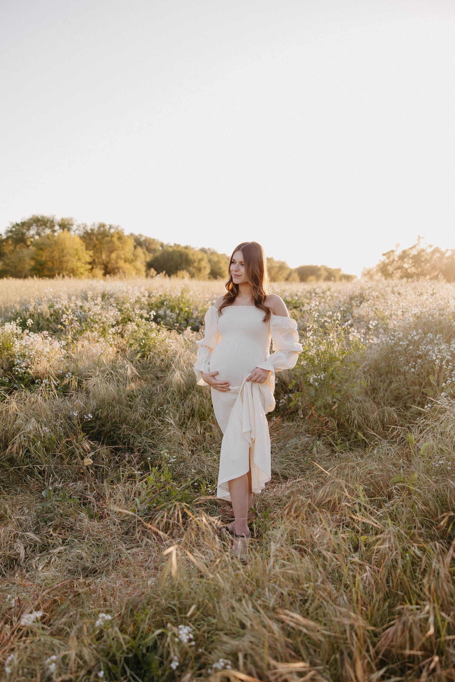 Dreamy maternity photo in white dress standing among tall grass and wildflowers.