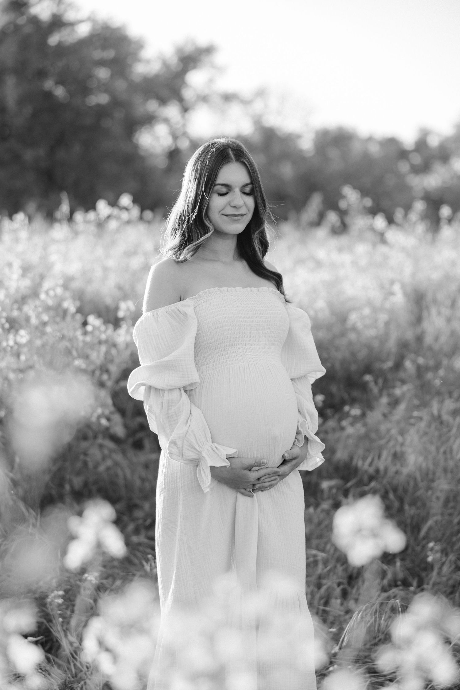 Black and white portrait of pregnant woman in flowing dress standing among wildflowers.
