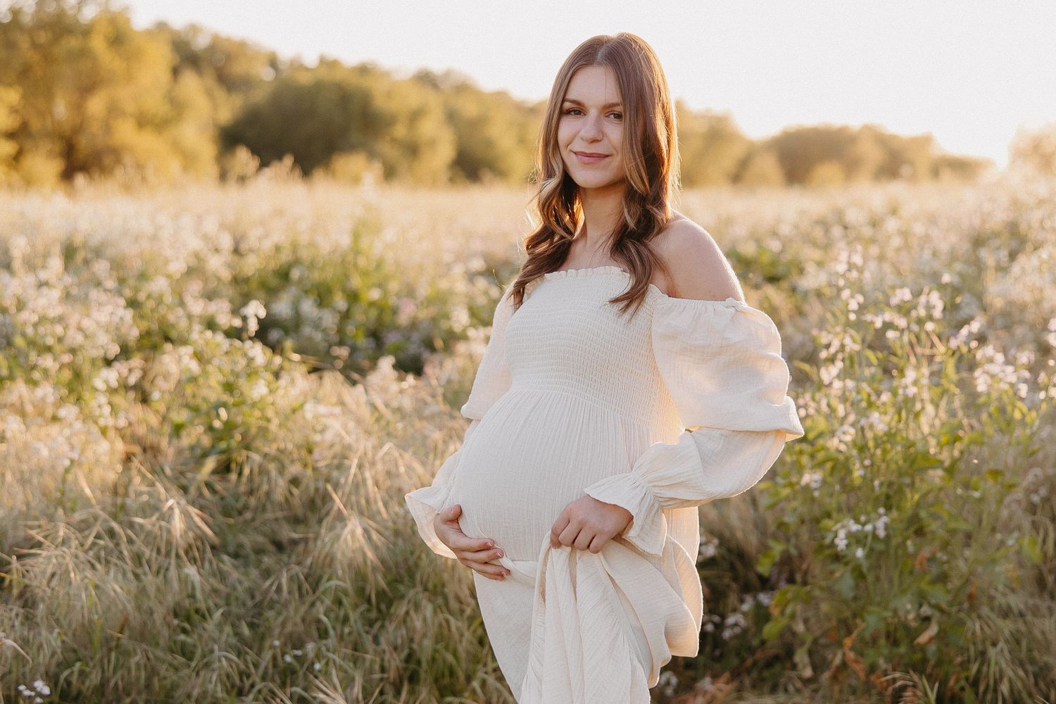 Maternity portrait in off-shoulder white dress amid wildflowers during golden hour.
