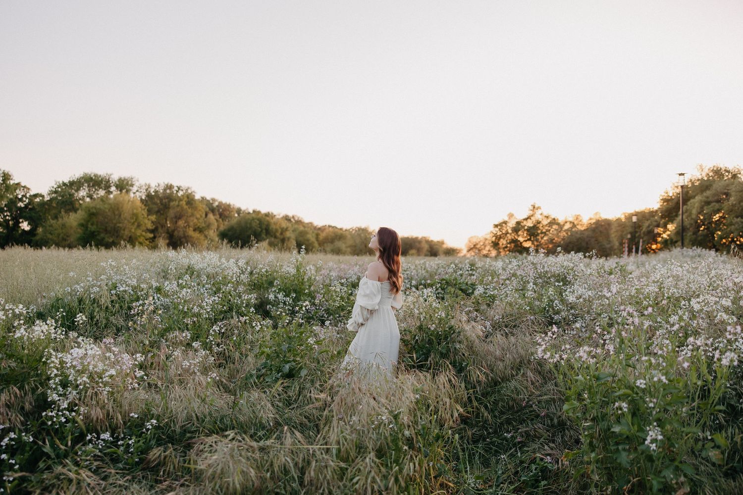 A dreamy figure in a white dress stands among wildflowers in a golden meadow at sunset.