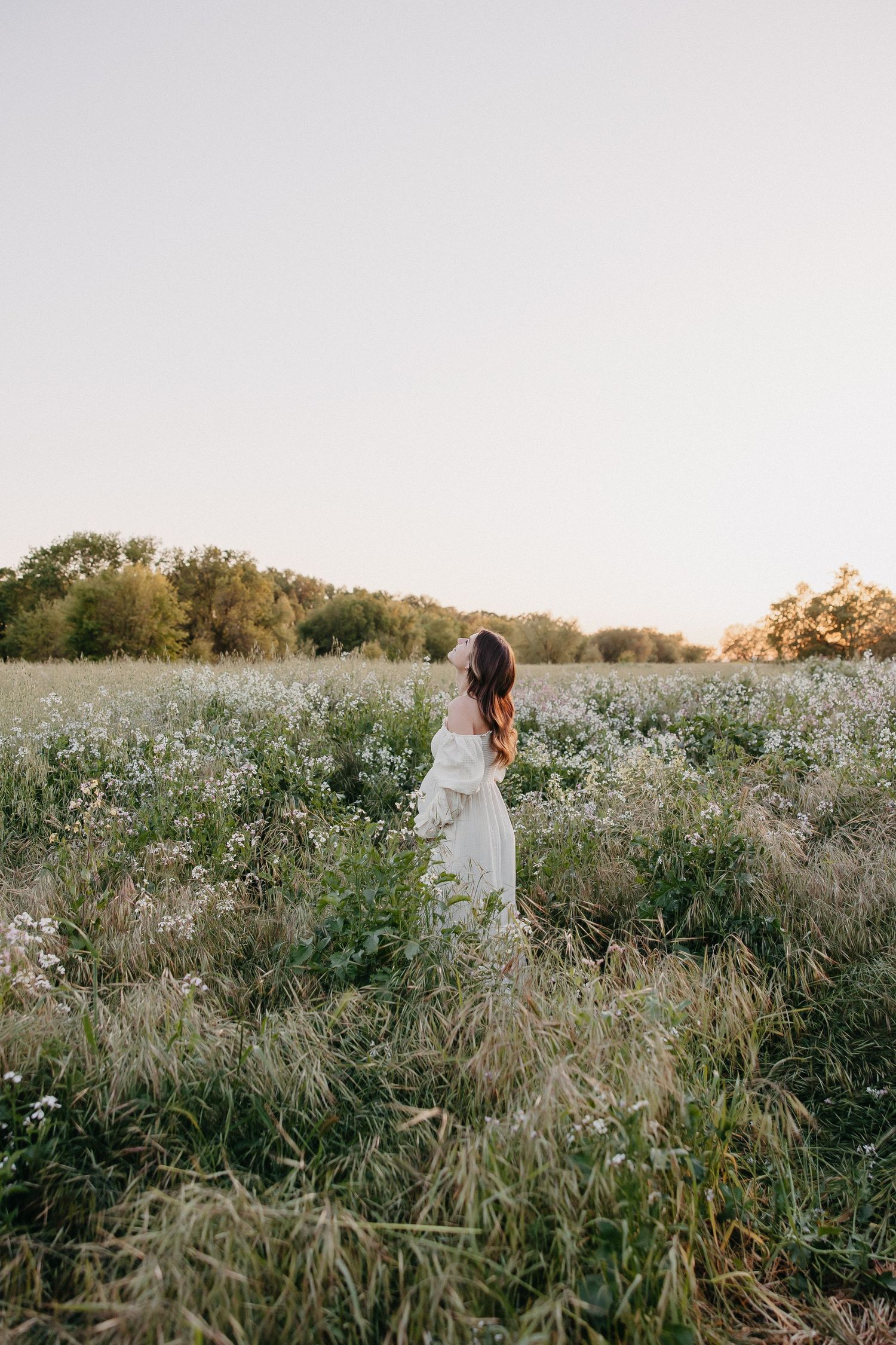 Back view of woman in white dress surrounded by wildflowers at sunset.