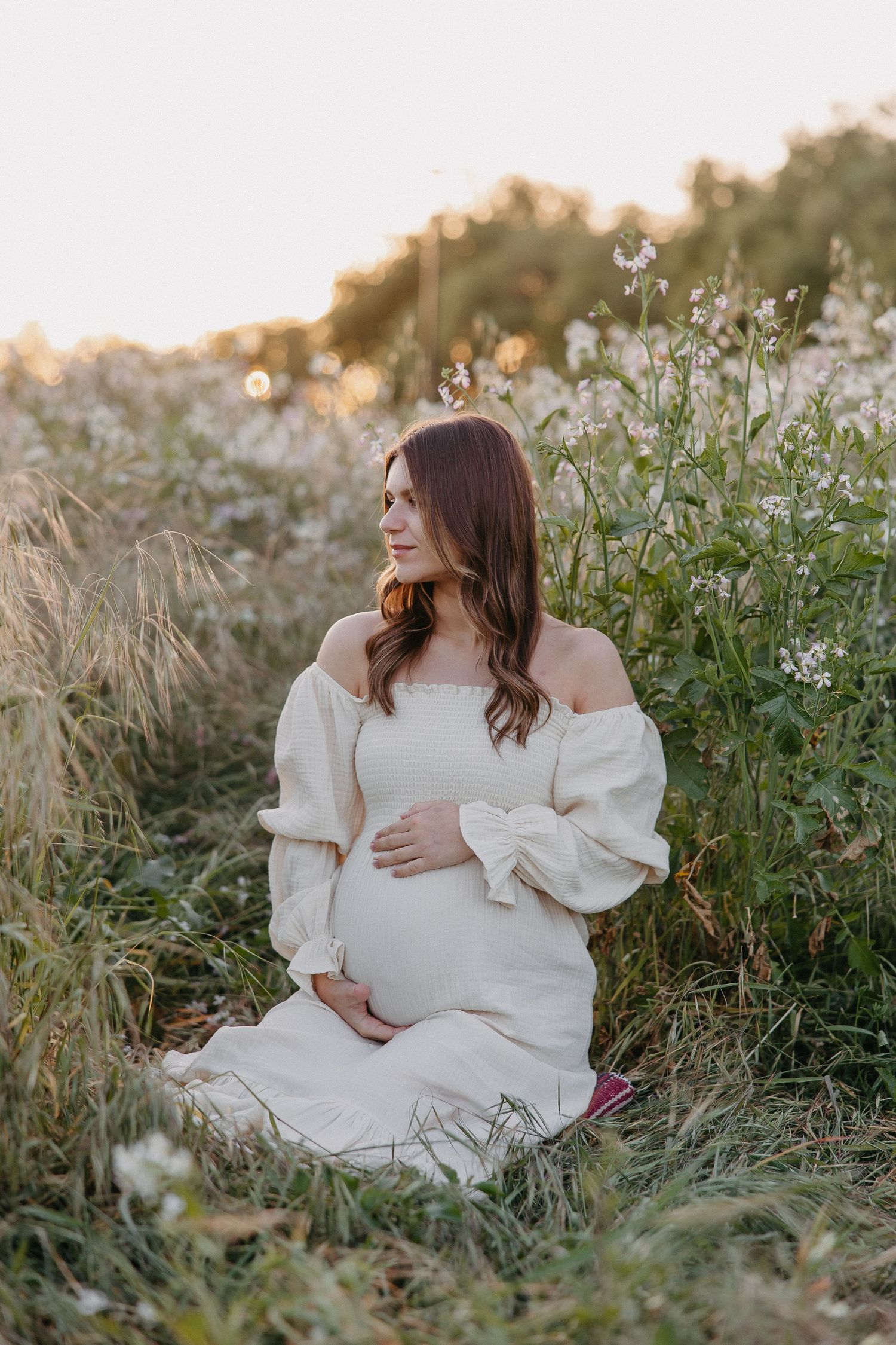 Serene maternity portrait seated in wildflower field wearing off-shoulder white gown.