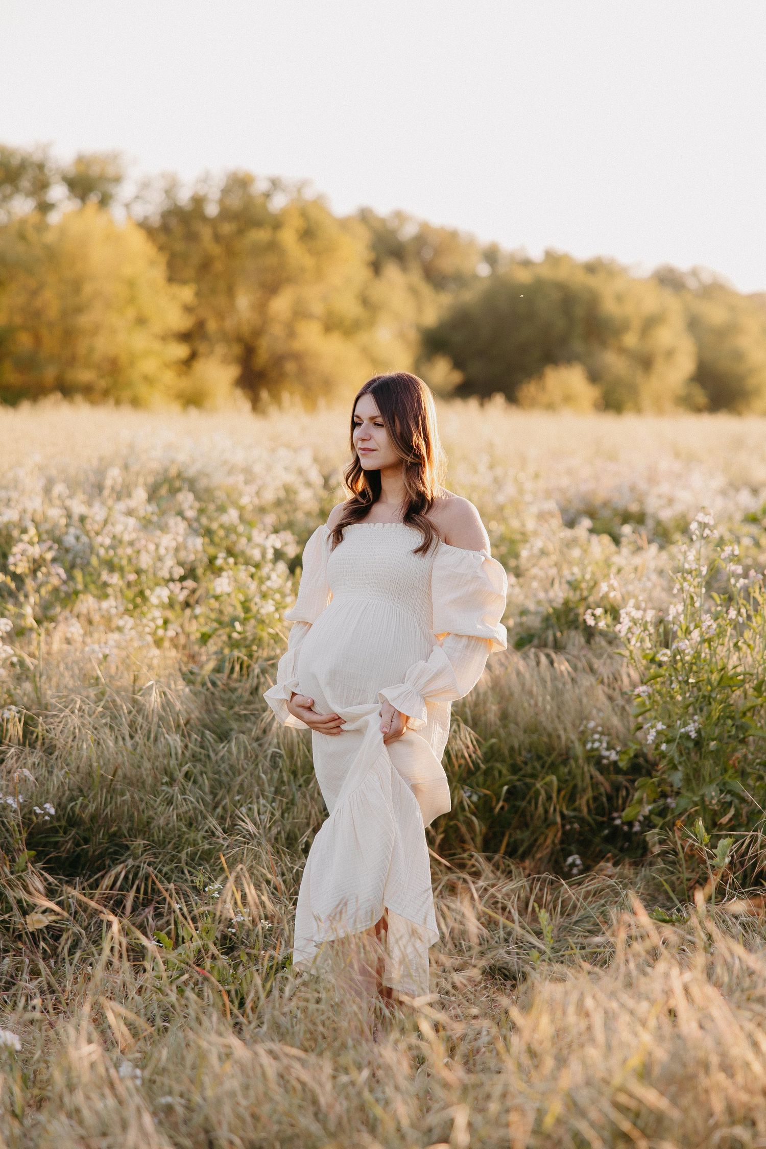 Expectant figure in white dress stands in golden field at sunset with flowing dress and long sleeves.