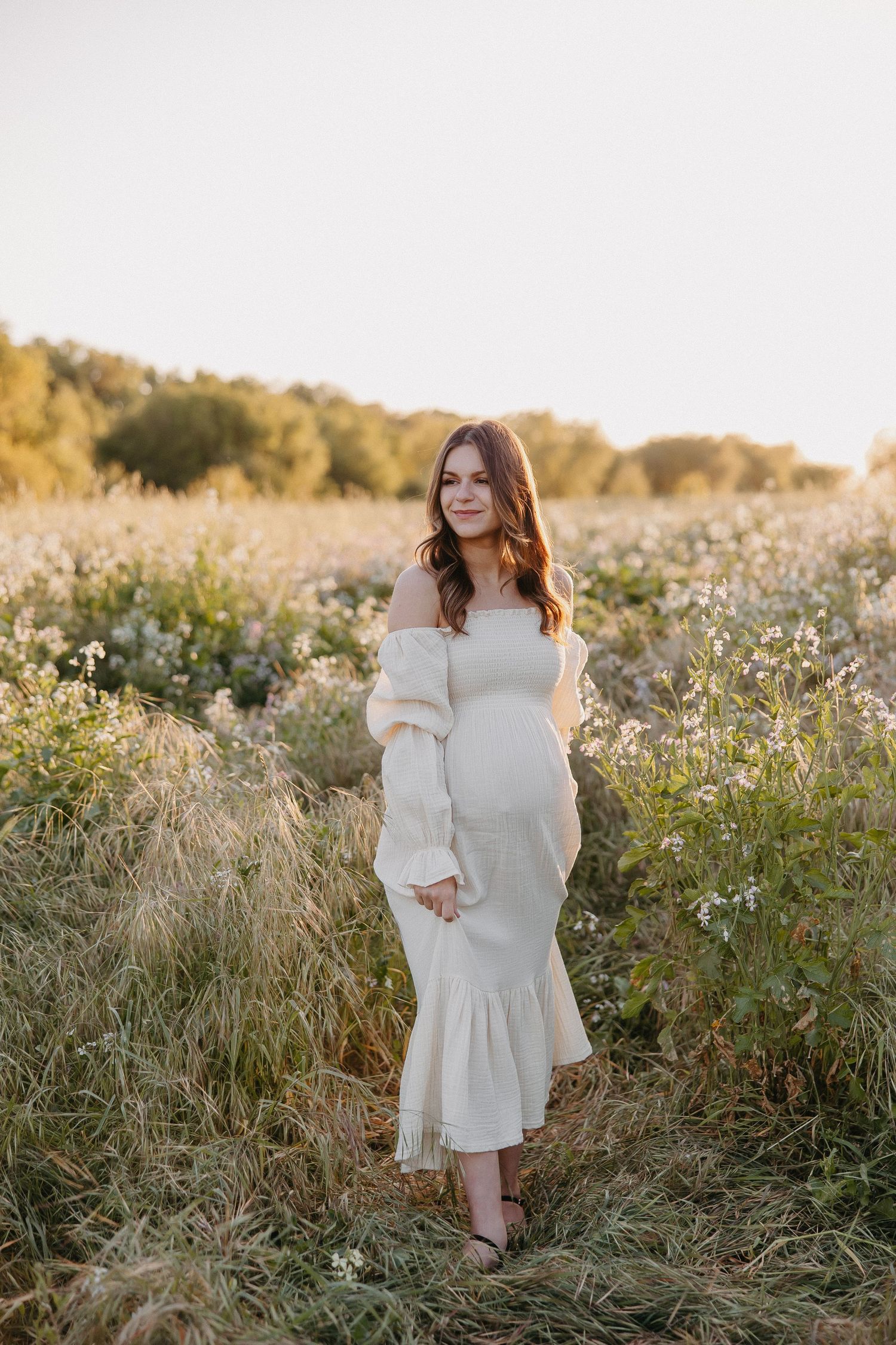 Elegant maternity pose in fitted white dress with ruffled hem amid wild grass.