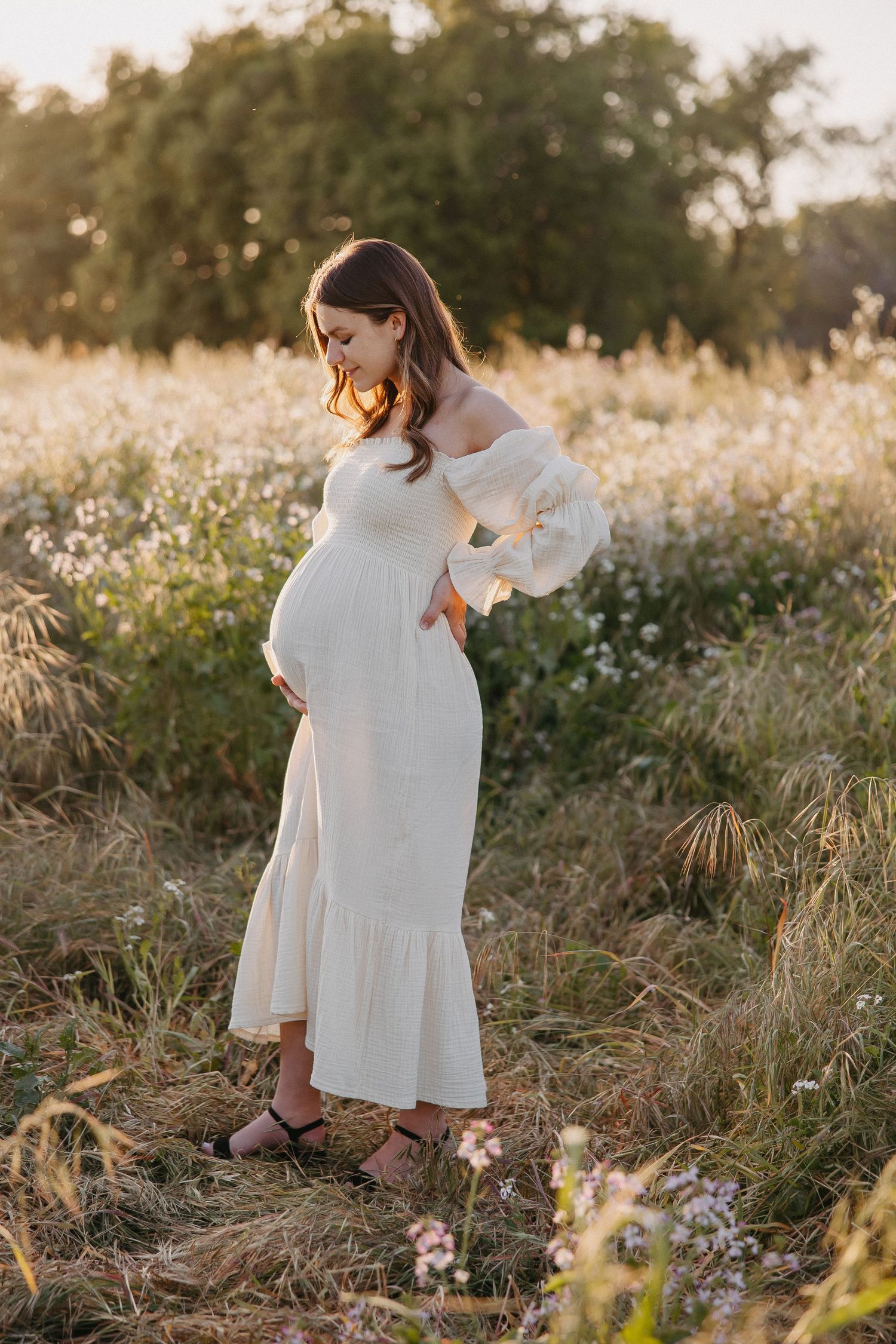 Side view of pregnant woman in white maxi dress standing amid wildflower meadow at sunset.