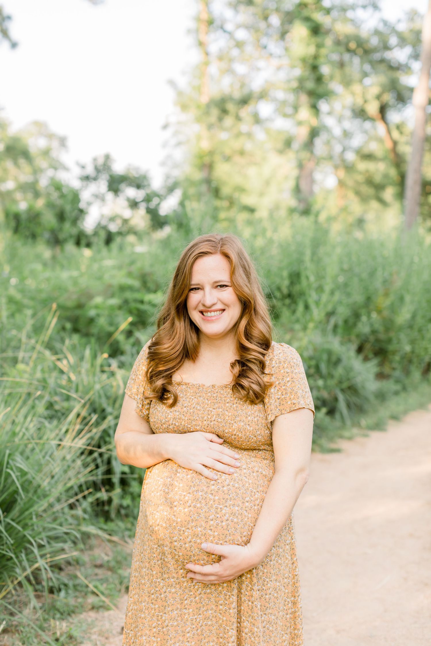 Woman in yellow dress smiles while holding baby bump on outdoor trail.