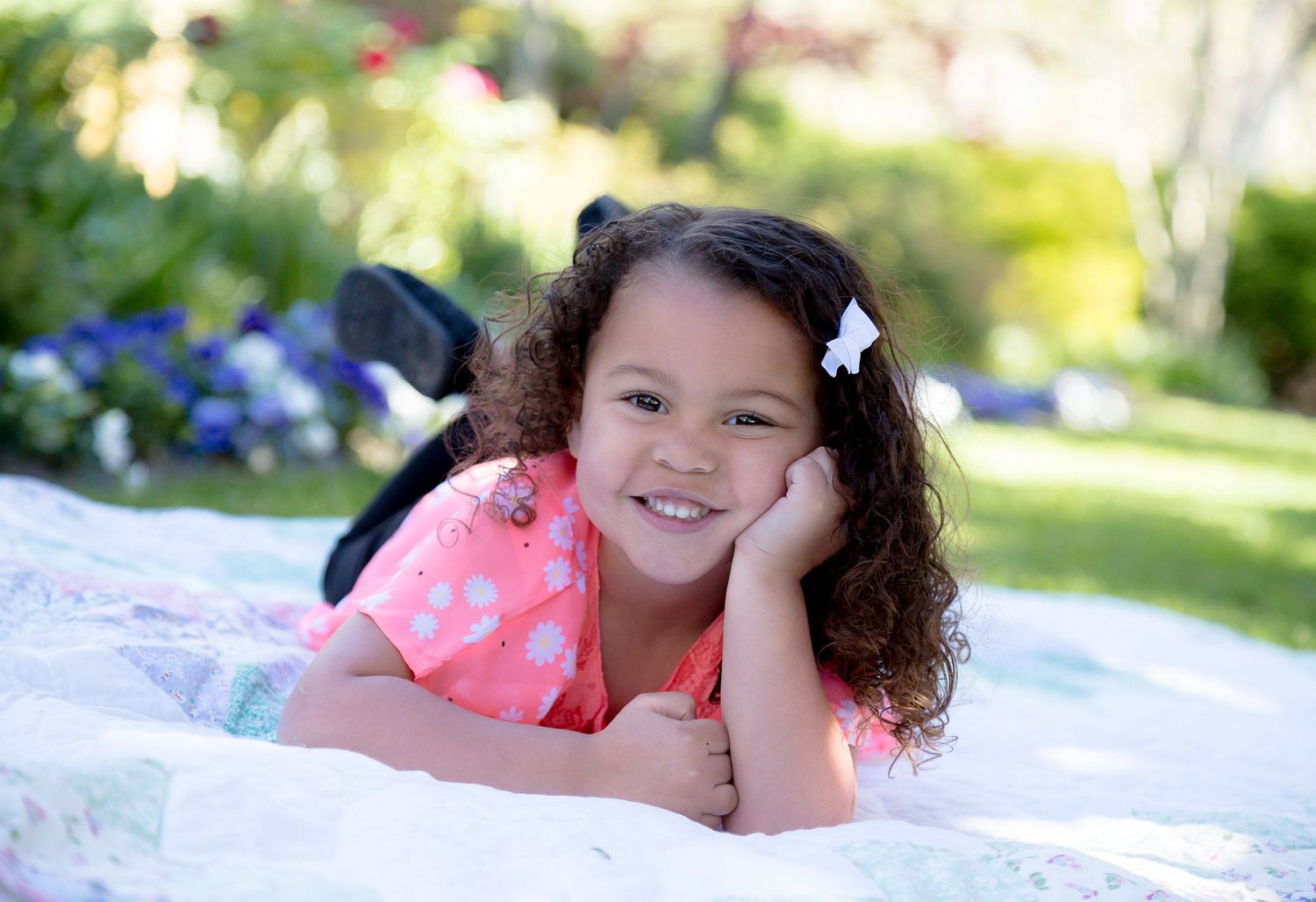 Young girl with curly hair in pink dress smiling while lying on blanket in garden.