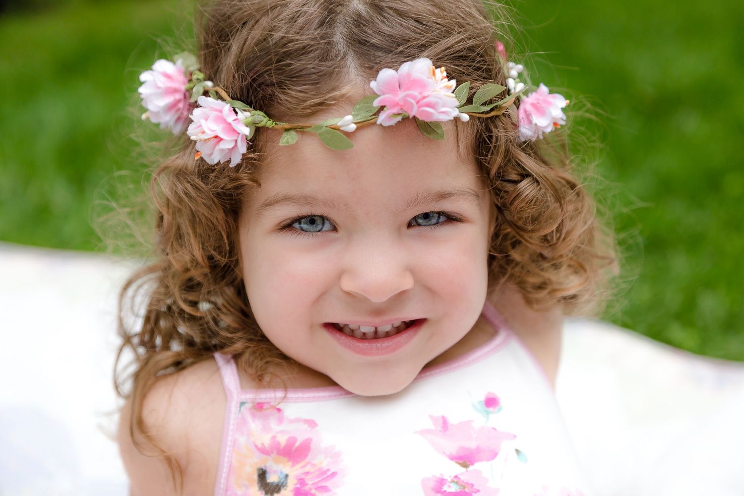 Adorable curly-haired little girl wearing pink flower crown smiling in sunny garden.