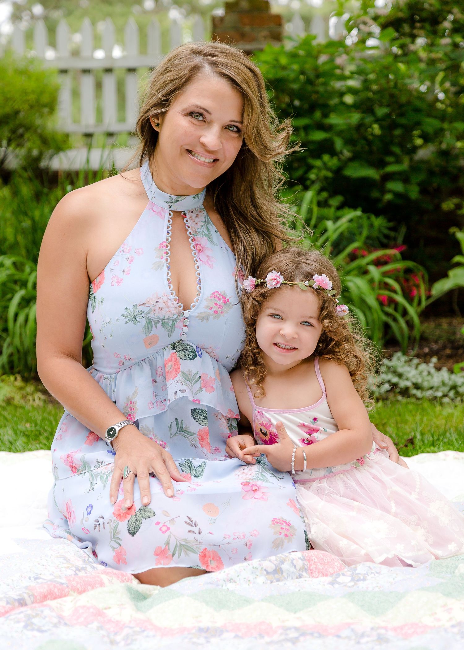 Smiling mother in floral blue dress sitting with curly-haired daughter in pink in garden.