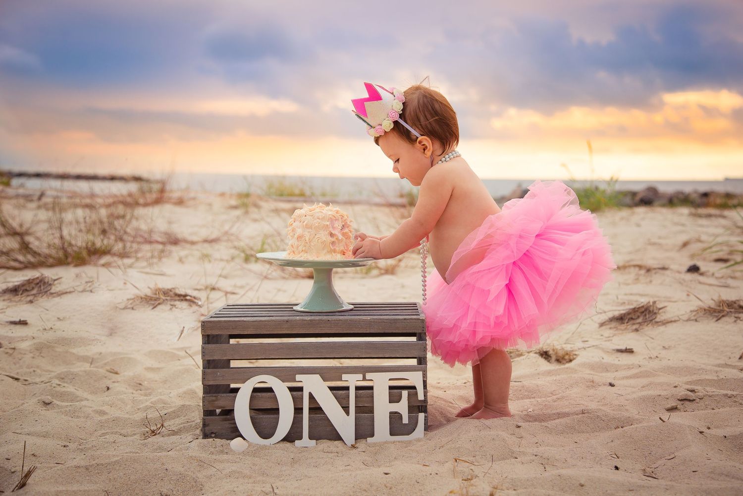 Baby girl in pink tutu smashing first birthday cake on beach with ONE sign at sunset.
