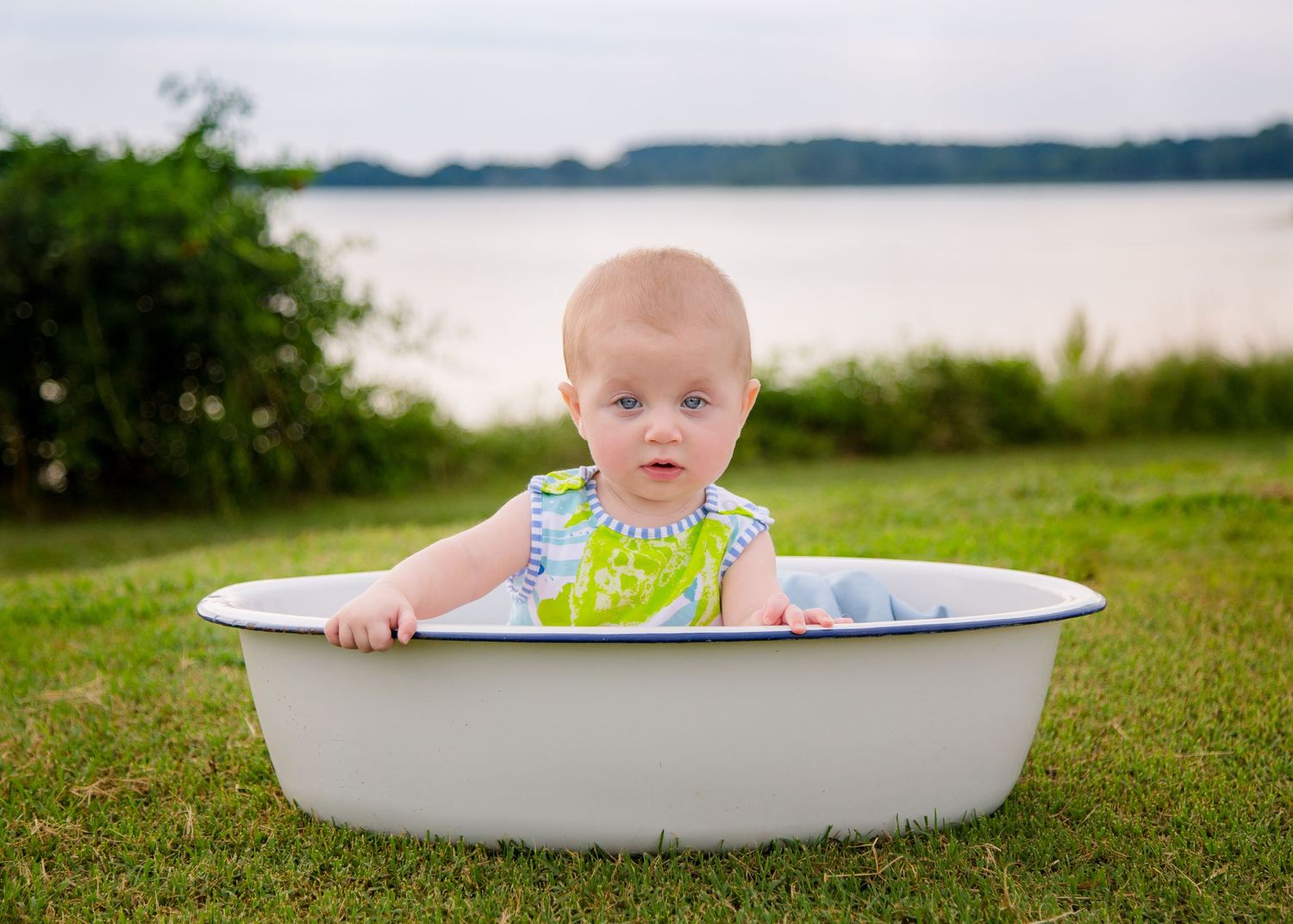 Adorable baby in floral outfit sitting in vintage enamel tub on green grass by lake.
