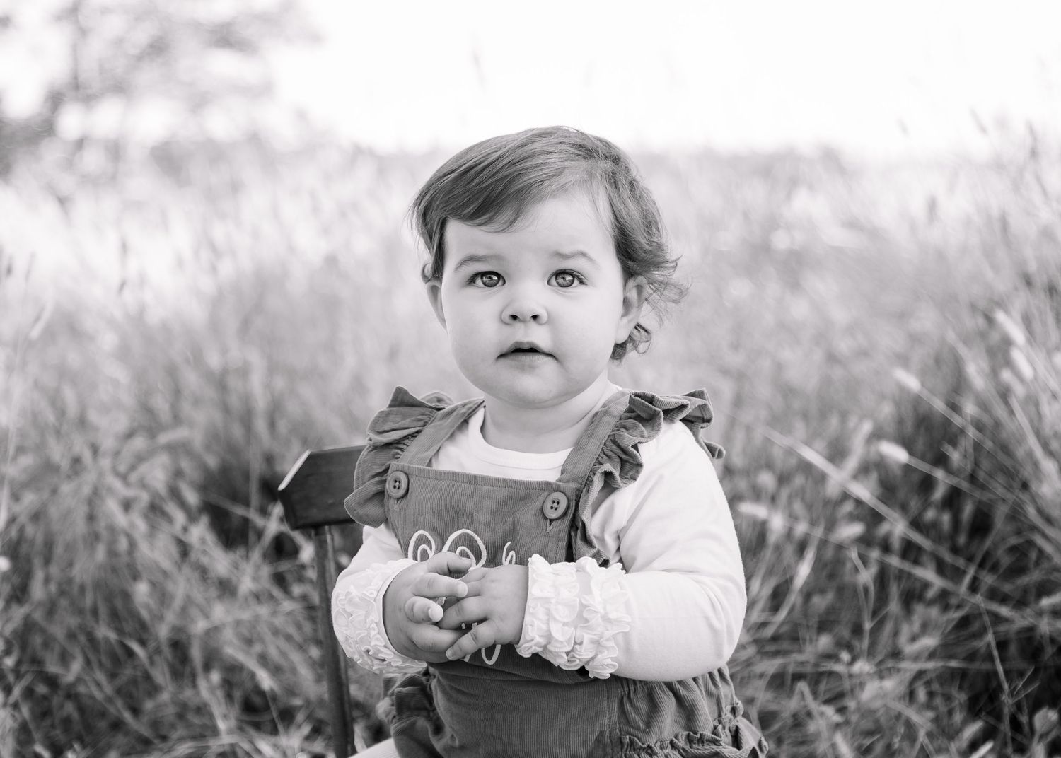 Black and white portrait of cute baby girl in overalls standing in tall grass field.