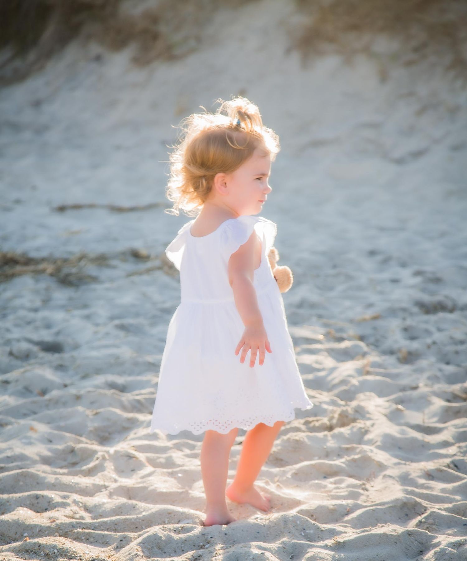 Toddler girl in white dress holding teddy bear standing barefoot on sandy beach.