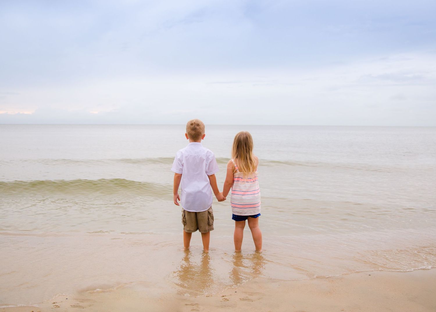 Two young children holding hands while standing at the ocean's edge on a cloudy beach day.