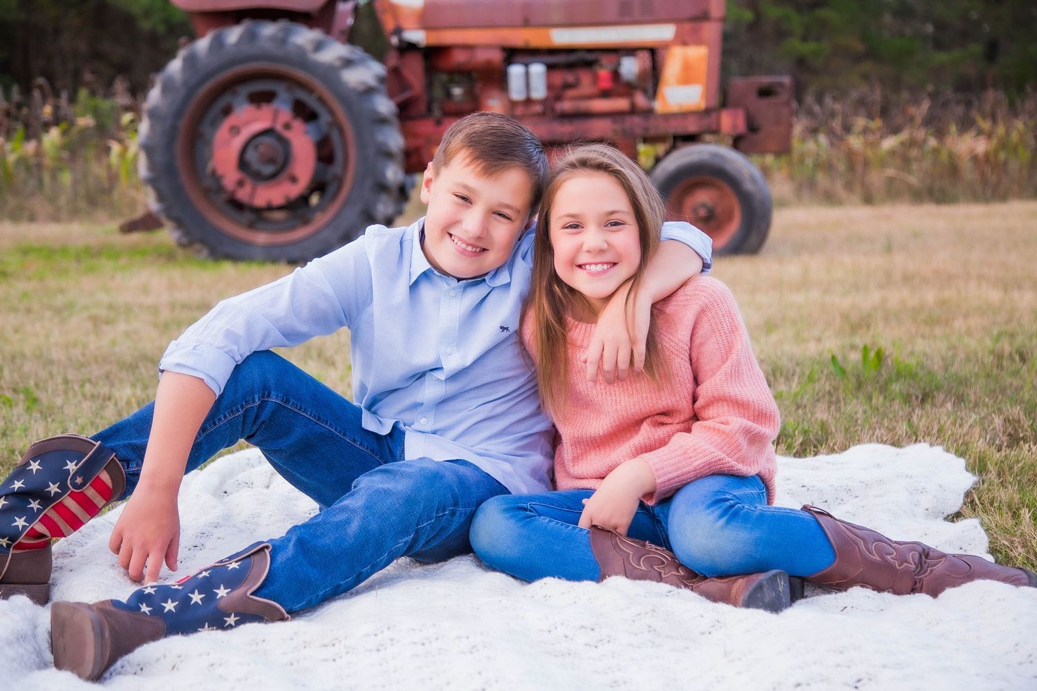 Smiling boy and girl siblings sitting together on a white blanket in a field near a red tractor.