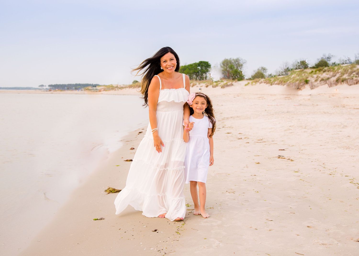 Mother and daughter in matching white dresses walking together on a sunny sandy beach.