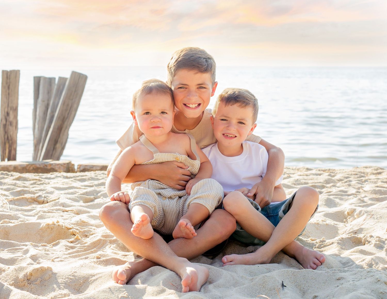 Older brother hugging two younger siblings on sandy beach at golden hour sunset.