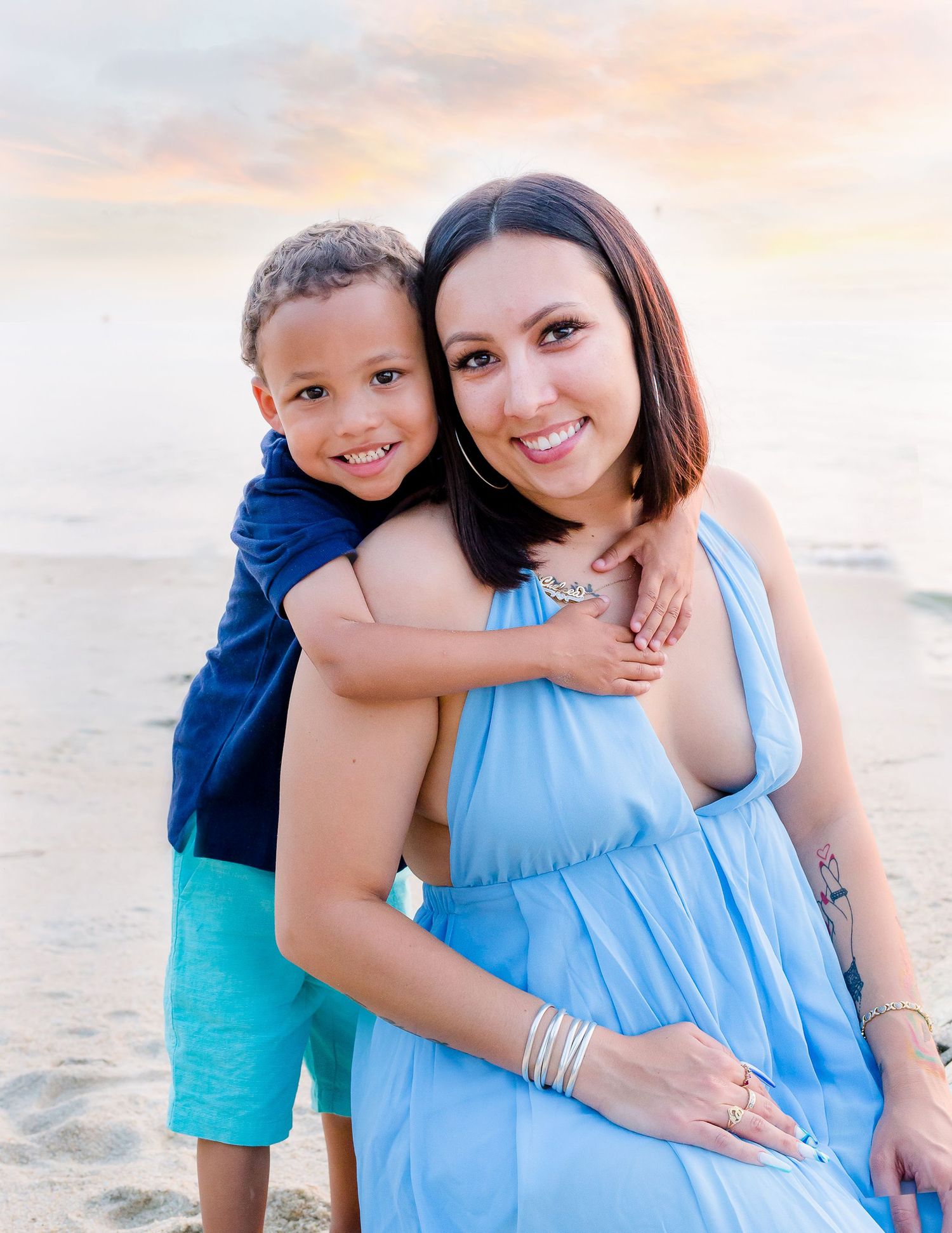 Pregnant mother in blue dress hugging her young son on a beach at sunset.