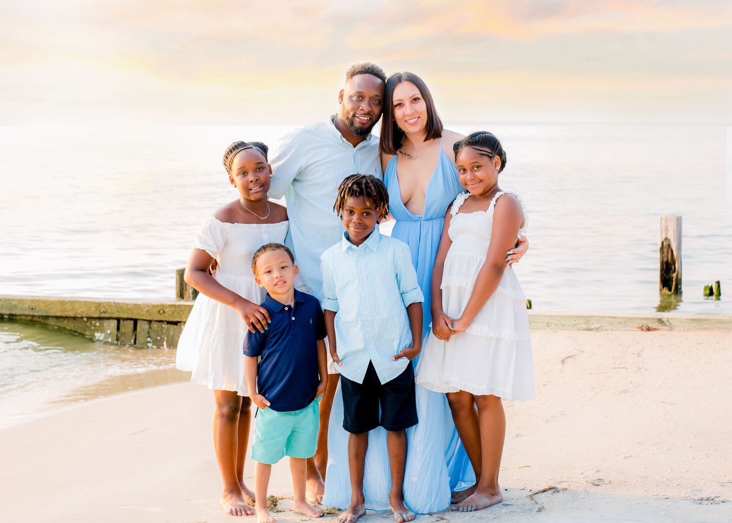 Joyful family of six posing together on a beach at sunset in coordinating white and blue outfits.